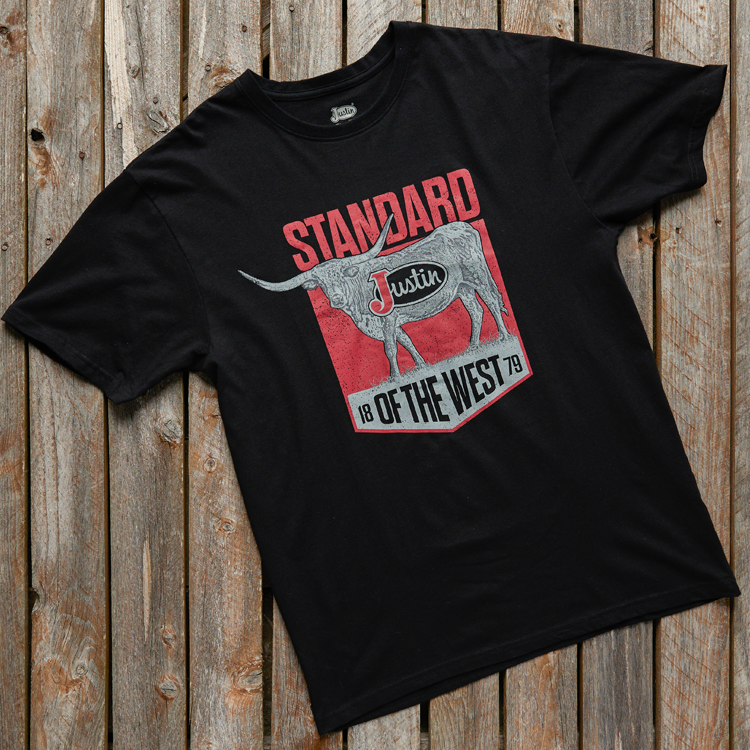 A black t-shirt that has a longhorn and says “Standard of the West” laying down on a wood background.